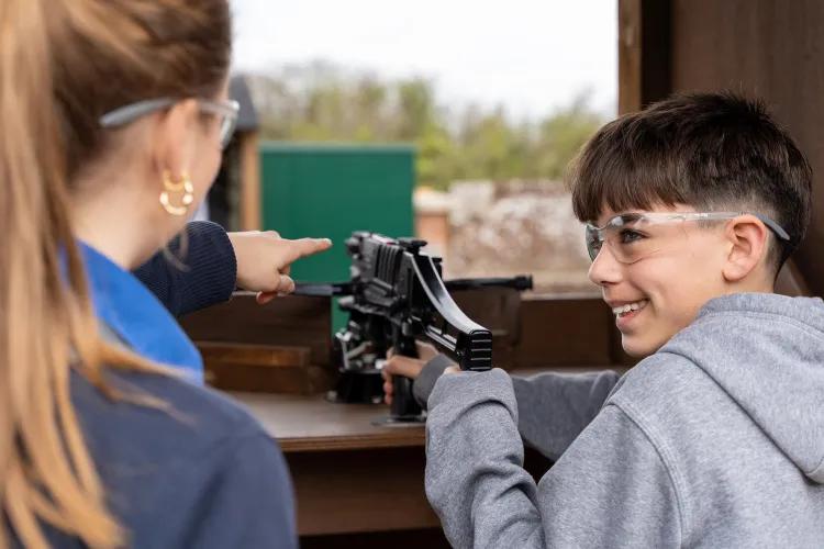 Two people at a crossbow range, one giving guidance and pointing as the other holds a mounted crossbow, outdoor backdrop