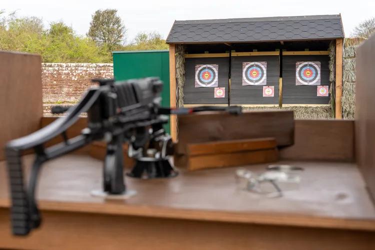 Crossbow fixed on a shooting stand in the foreground, with three archery-style targets aligned beneath a small shelter