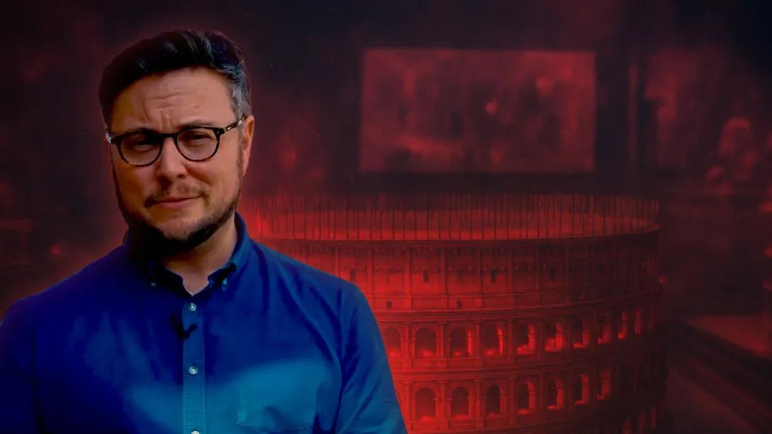 a young man in blue shirt with short brown hair and beard and glasses stands in front of colosseum motifs